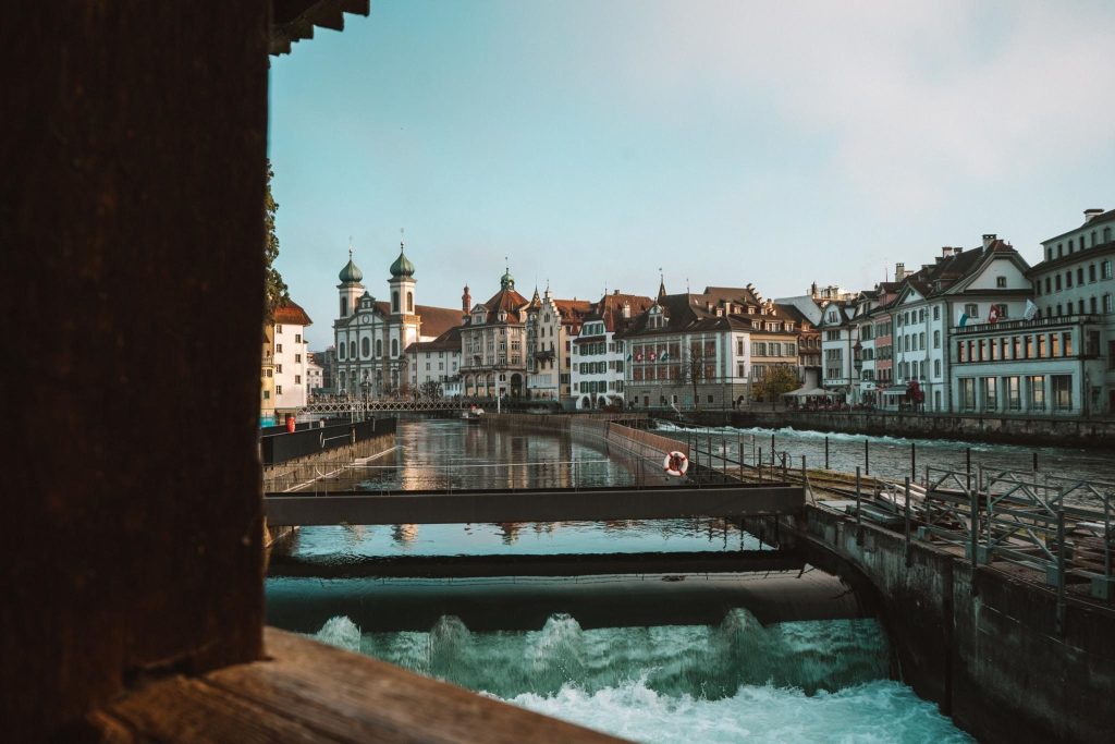 Een schilderachtig uitzicht op een Europese stad met historische gebouwen, een prominente kerk met twee groene koepels, een rivier met kleine watervallen en een houten brug op de voorgrond onder een heldere hemel.