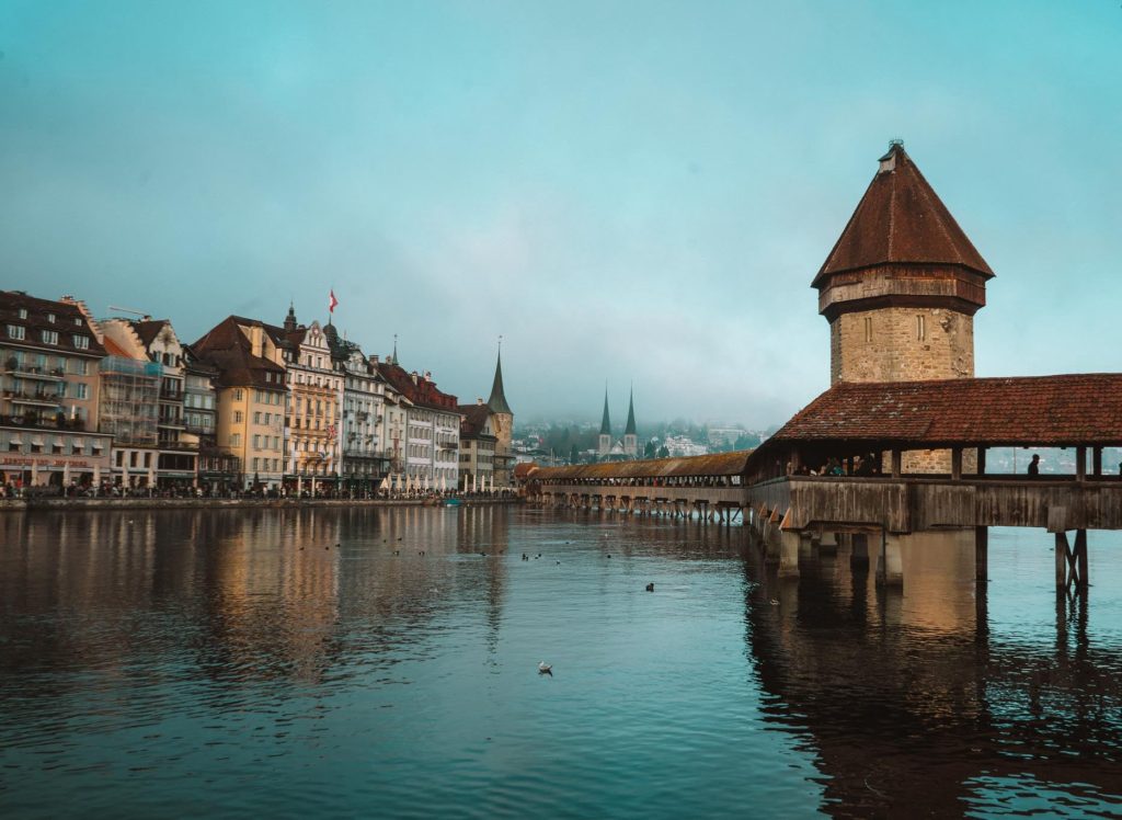 Een historische houten brug met een stenen toren strekt zich uit over kalm water in een Europese stad, met oude gebouwen langs de oever en kerktorens zichtbaar op de achtergrond onder een blauwe hemel.
