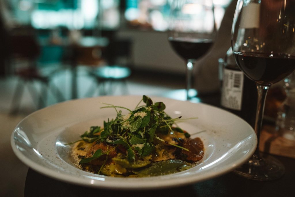 Een close-up van een witte kom gevuld met kleurrijke ravioli pasta, gegarneerd met verse groenten en geraspte kaas, met twee glazen rode wijn op een tafel in een zacht verlicht restaurant.