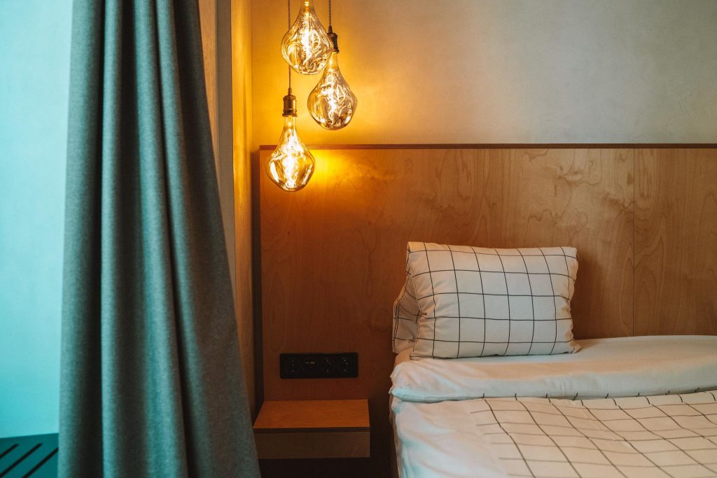 Een gezellige slaapkamerhoek met een bed met een wit kussen en deken met rasterpatroon, warme hanglampen, een houten hoofdeinde en een grijs gordijn dat gedeeltelijk aan de zijkant is dichtgetrokken.