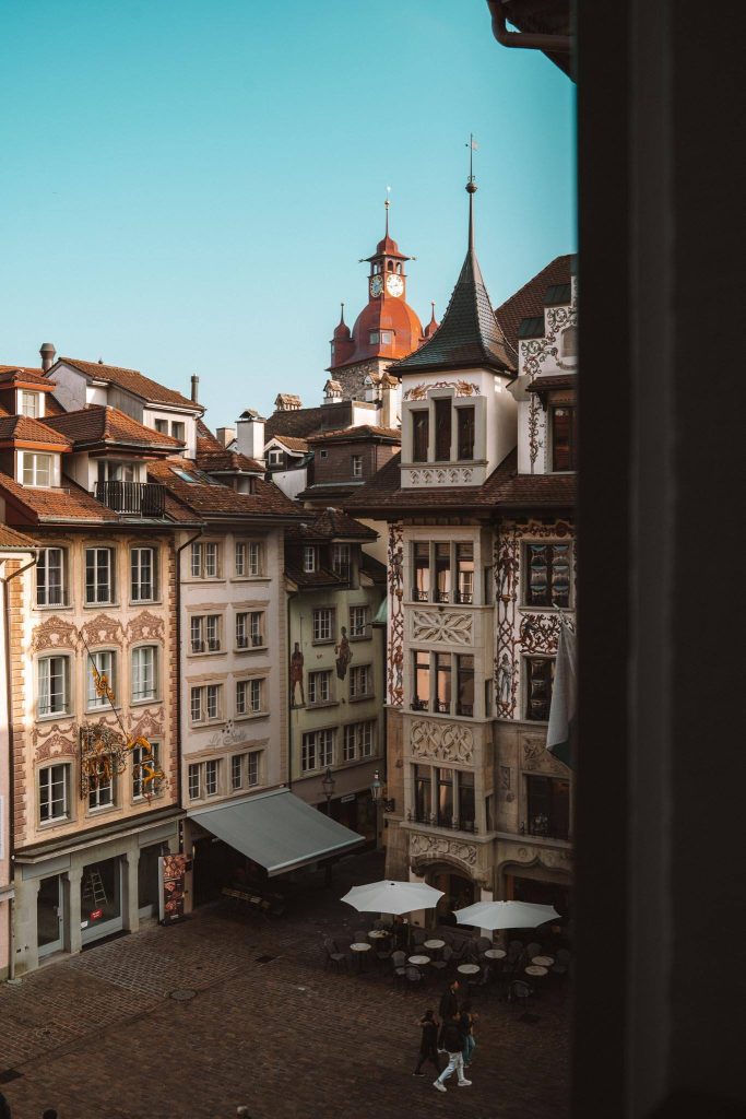 Een charmant Europees plein met sierlijke, historische gebouwen, cafétafels in de open lucht en een paar wandelende mensen. Op de achtergrond steekt een rode klokkentoren boven de daken uit tegen een strakblauwe lucht.