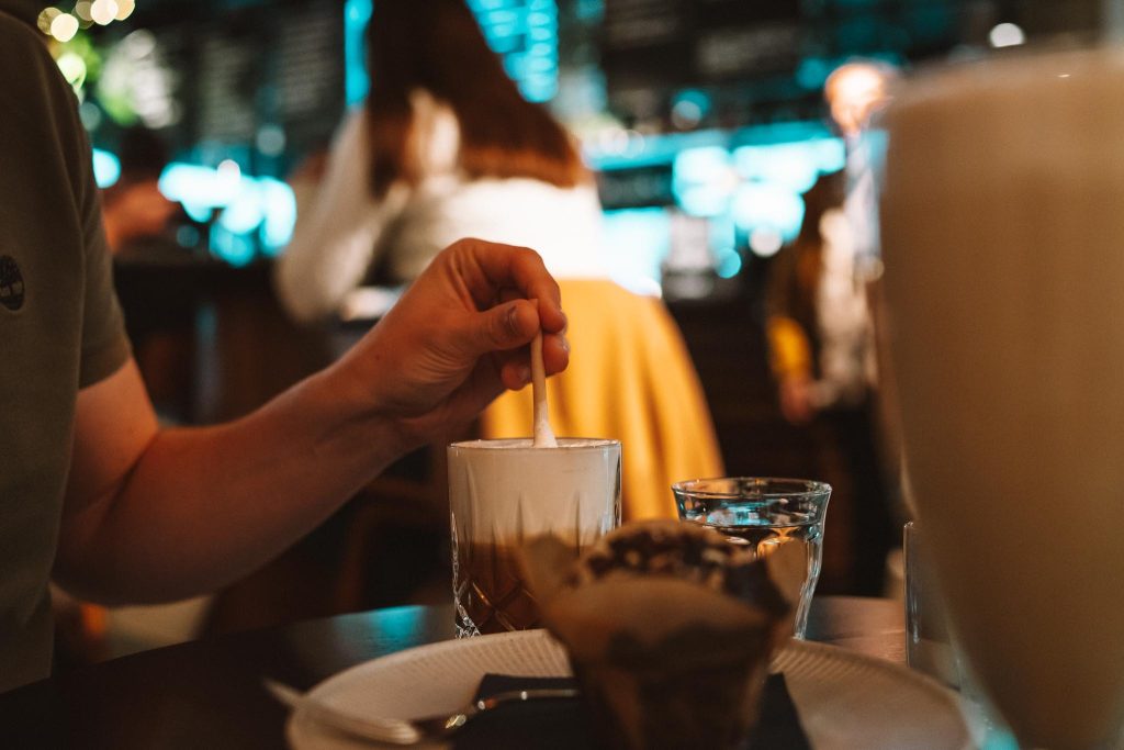 De hand van een persoon roert een romig drankje met een rietje aan een cafétafel, waar ook een glas water en een muffin staan. Op de wazige achtergrond zitten mensen te socializen.