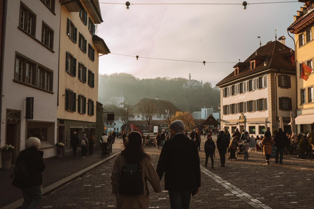 Een stel loopt hand in hand door een geplaveide straat met historische gebouwen, met veel mensen voor zich en heuvels zichtbaar op de achtergrond onder een bewolkte hemel.