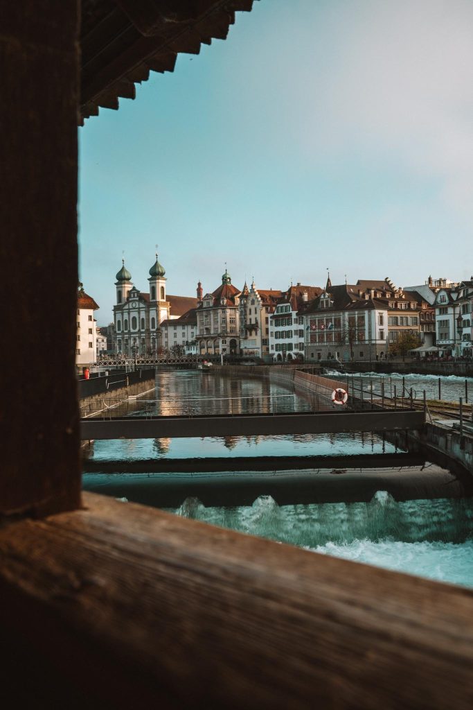 Gezicht op een Europese rivierstad met historische gebouwen, een kerk met groene koepels, bruggen over rustig water en weerspiegelingen onder een helderblauwe hemel, omlijst door een houten structuur.
