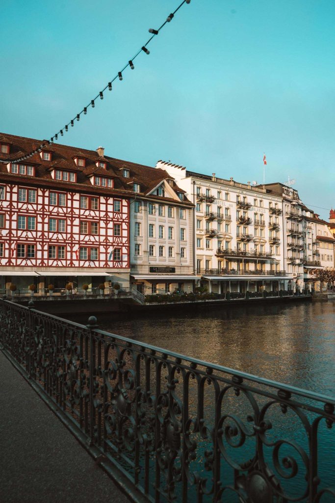 Een rij historische gebouwen met decoratieve gevels staat langs een rivier, gezien vanachter een zwarte versierde reling onder strijklicht, onder een strakblauwe hemel.