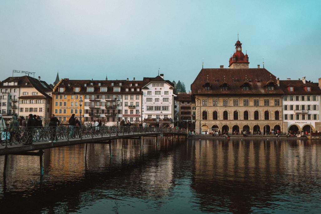Een schilderachtig uitzicht op historische gebouwen langs de waterkant in Luzern, Zwitserland, met mensen die over een metalen loopbrug over het kalme water onder een blauwe hemel lopen.