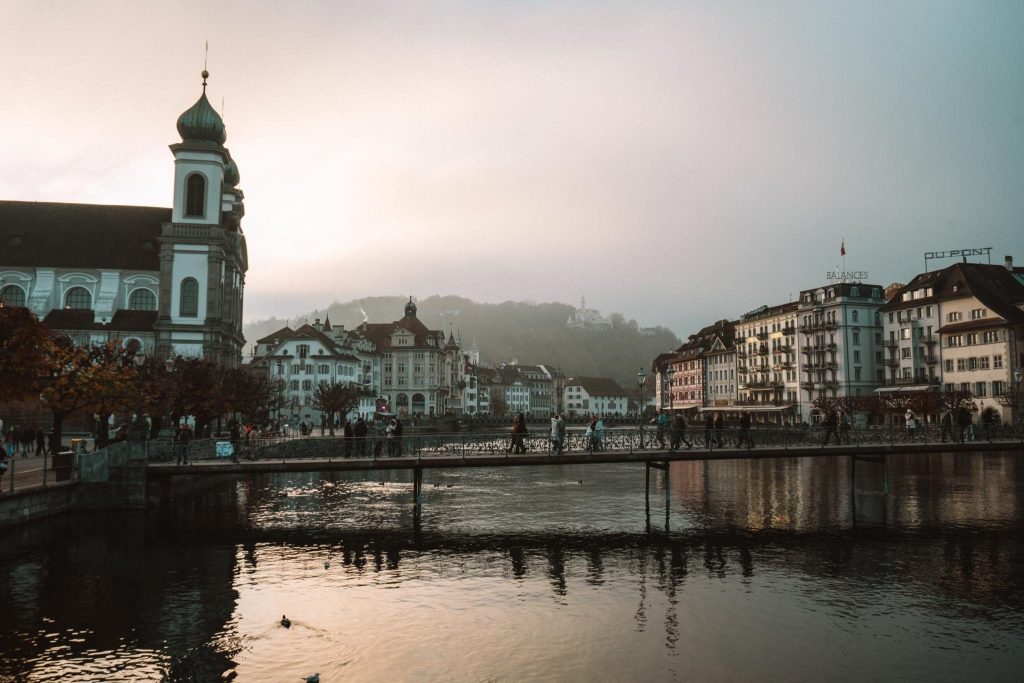 Een schilderachtig uitzicht op een Europese stad bij zonsondergang, met historische gebouwen en een kerk aan een rustige rivier. Mensen steken een smalle brug over het water over en op de achtergrond zijn mistige heuvels zichtbaar.
