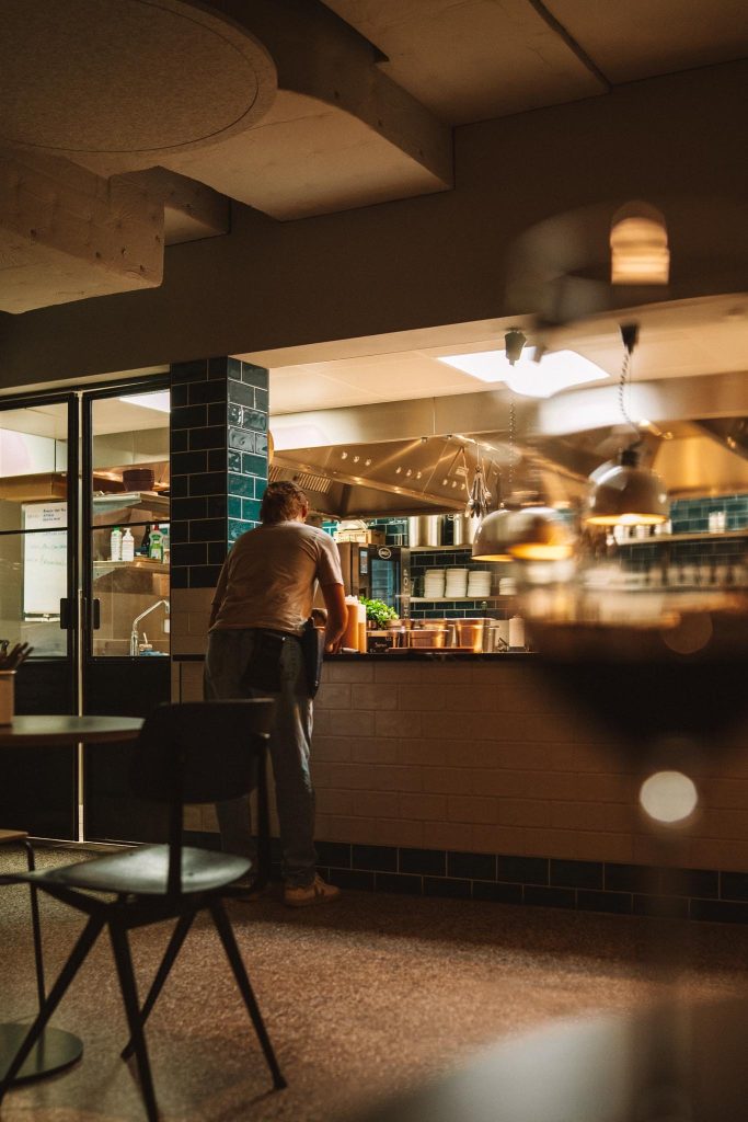 Staande en werkende persoon aan een aanrecht in een warm verlicht restaurant, met planken, keukengerei en groen zichtbaar. Een tafel en stoel zijn op de voorgrond, onscherp.