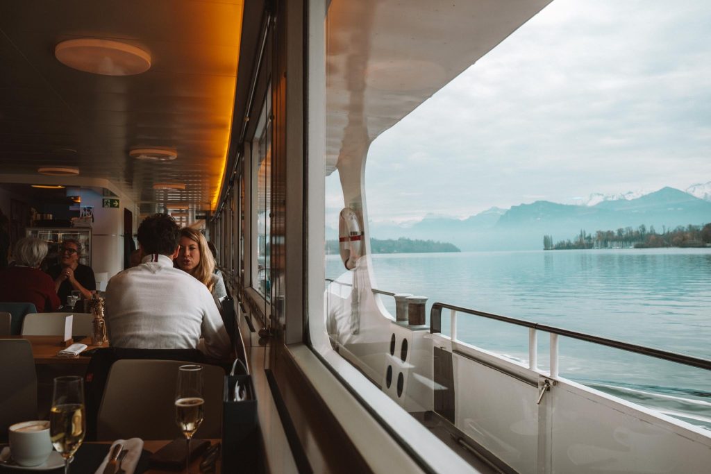 Mensen zitten aan tafels in een boot met grote ramen en kijken uit op een rustig meer omringd door mistige bergen. Op tafel staan een glas wijn en water. Het tafereel is vredig en ontspannen.
