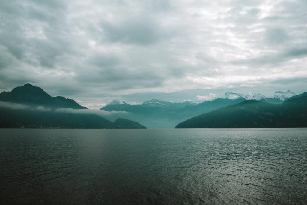 Een kalm meer onder een bewolkte hemel, omringd door bergen met besneeuwde toppen in de verte en mist die over het water hangt.