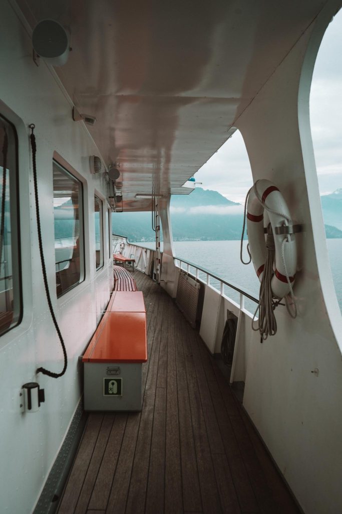 Een houten dek op een veerboot of schip met oranje banken, reddingsringen aan de muur en grote ramen, met uitzicht op een watermassa en verre mistige bergen onder een bewolkte hemel.