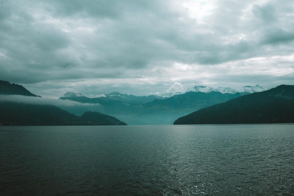 Een rustig meer met donker water strekt zich uit in de richting van verre, mistige bergen. Besneeuwde toppen en beboste heuvels zijn zichtbaar onder een bewolkte lucht.