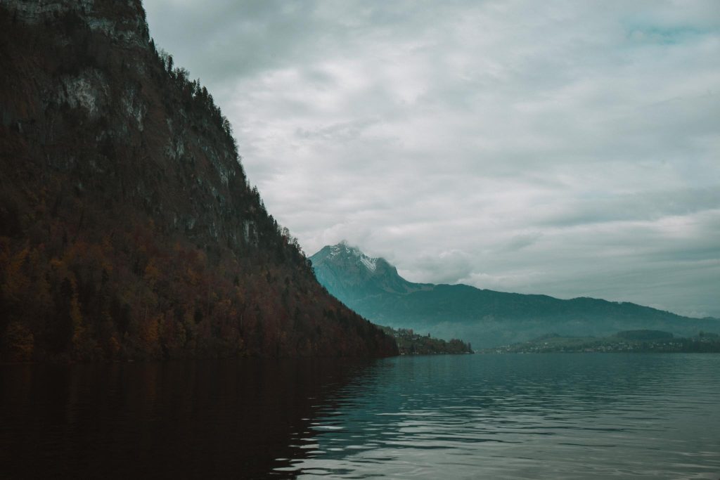Een kalm meer omzoomd door een beboste berghelling aan de linkerkant, met in de verte een besneeuwde top onder een bewolkte hemel. Het water weerspiegelt het landschap en creëert een serene en rustige sfeer.