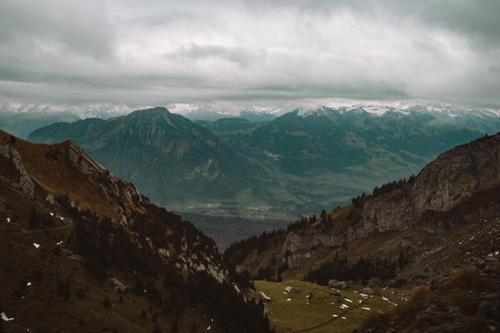 Een bewolkte hemel hangt boven een schilderachtig berglandschap met ruige kliffen, verspreide sneeuwvlokken, groene valleien en toppen in de verte die gedeeltelijk in mist gehuld zijn.