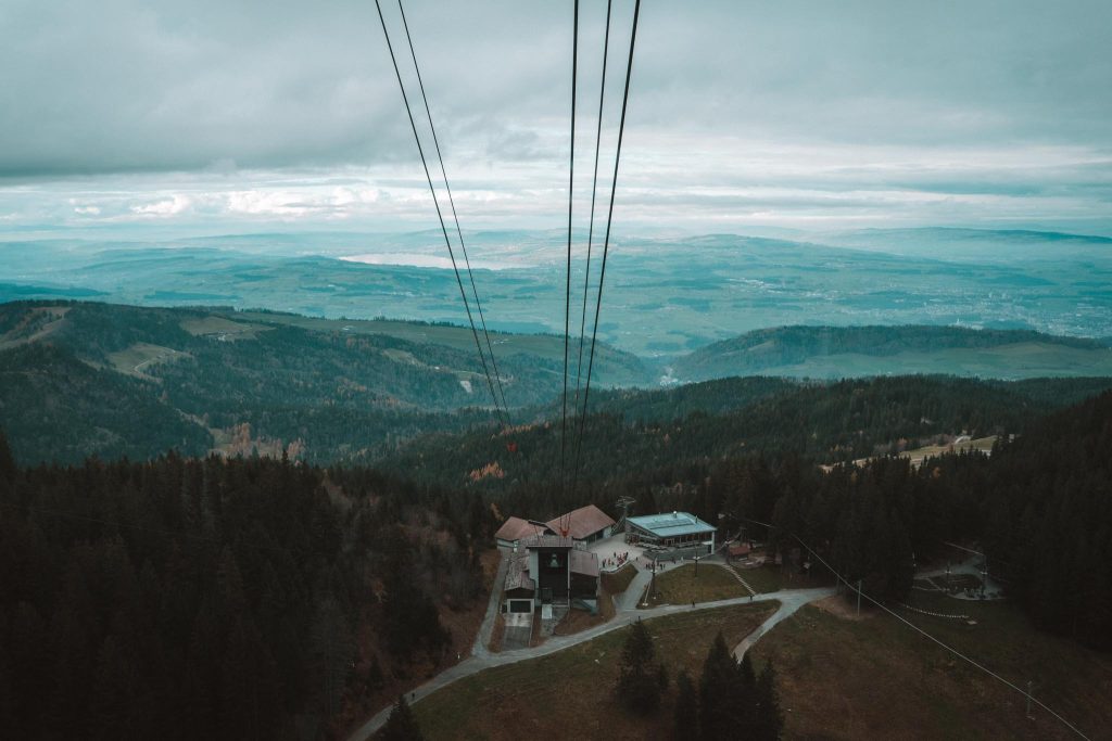 Een schilderachtige luchtfoto van een kabelbaanstation omringd door dichte bossen en glooiende heuvels, met kabels die zich uitstrekken naar de bewolkte hemel en een uitgestrekt landschap zichtbaar in de verte.