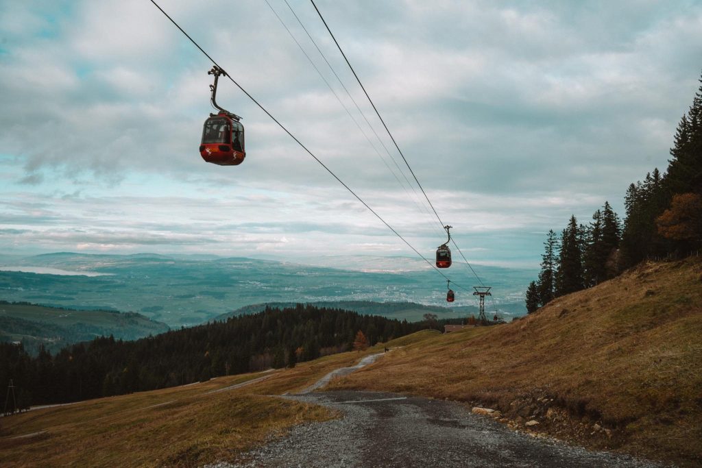 Rode kabelbanen rijden over grashellingen met verspreide bomen een berg op. Onder de kabels loopt een grindpad en op de achtergrond strekt zich een schilderachtig landschap uit onder een bewolkte hemel.