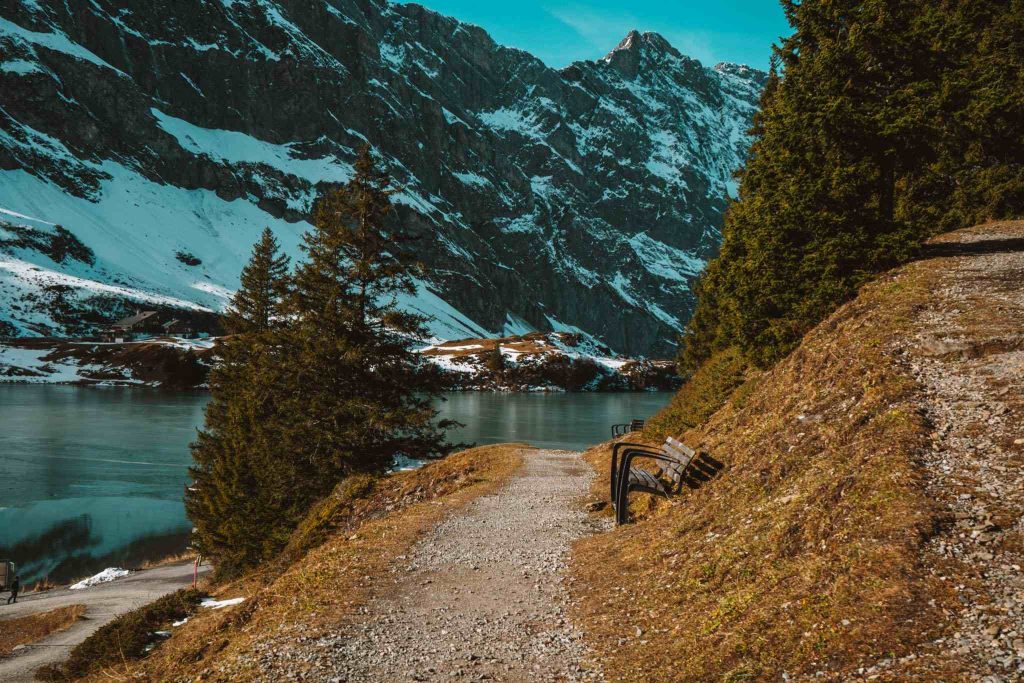Een grindpad met een houten bankje kronkelt langs een heuvel, met uitzicht op een kalm meer en besneeuwde bergen met altijd groene bomen onder een blauwe hemel.