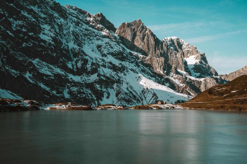 Een sereen meer met stilstaand water weerspiegelt besneeuwde grillige bergen onder een helderblauwe hemel. Op de achtergrond rijzen steile rotsen op en langs de oever liggen stukken sneeuw en gras.