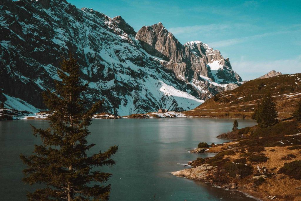 Een sereen bergmeer met helder water, omringd door groenblijvende bomen. Op de achtergrond rijzen besneeuwde rotsbergen op onder een blauwe hemel, waardoor een vredig, natuurlijk landschap ontstaat.