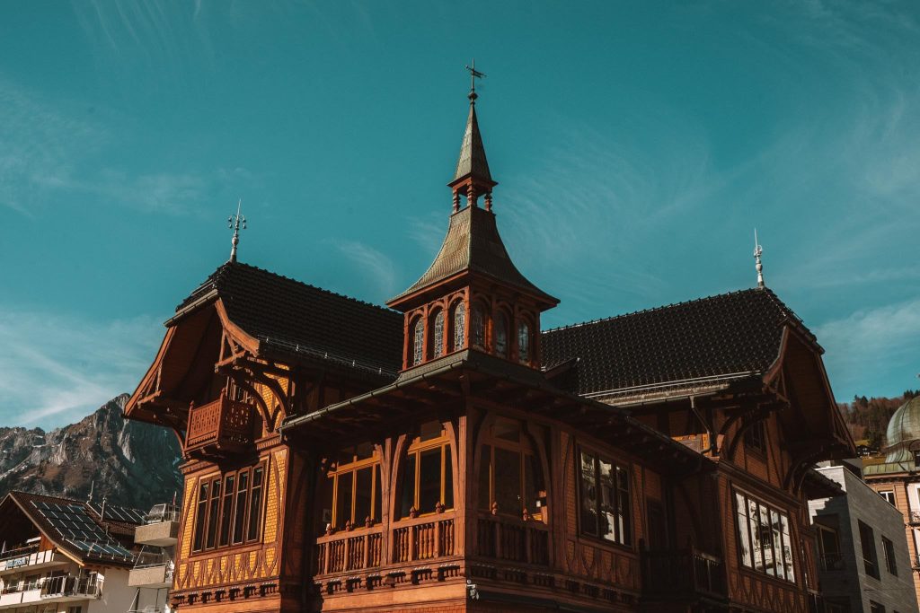 Een houten gebouw met sierlijke architectuur, steile daken en een centrale torenspits staat tegen een helderblauwe lucht, met bergen zichtbaar op de achtergrond.