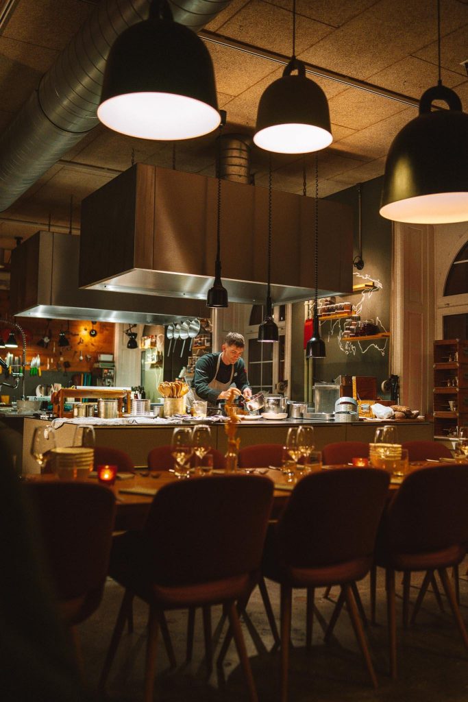 Een chef-kok bereidt eten in een moderne, warm verlichte restaurantkeuken. Op de voorgrond is een eettafel gedekt met wijnglazen en borden, terwijl hangende lampen de gezellige sfeer verlichten.
