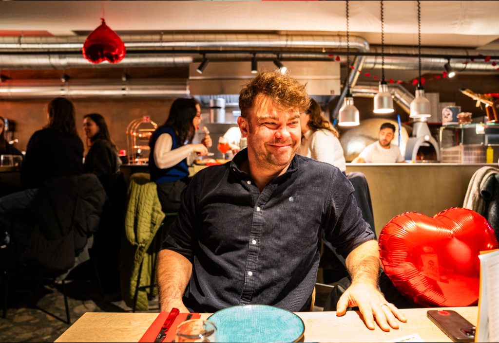 Een lachende man zit aan een restauranttafel in Leuven met een rode hartvormige ballon naast hem. Andere eters en rode hartversieringen zijn zichtbaar in de levendige, warm verlichte achtergrond - een viering van de liefde in een van de beste restaurants van de stad voor 2026.