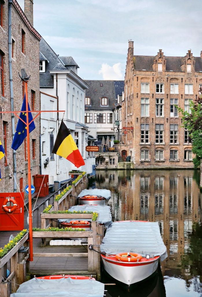 Kanaalscène in Brugge, België, met aangemeerde boten, Belgische en EU-vlaggen, bakstenen gebouwen en weerspiegelingen in het water. Historische architectuur en groen onder een halfbewolkte hemel roepen de charme op van enkele van de mooiste casino's in België.