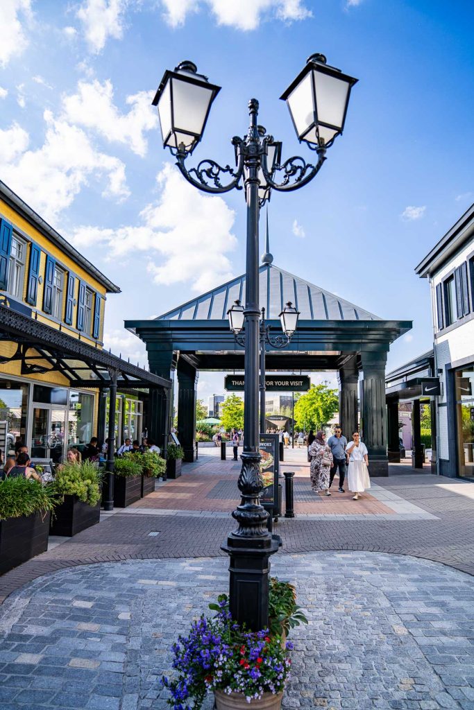 Op de kasseien staat een zwarte, sierlijke straatlantaarn, met bloempotten aan de voet. Winkelaars lopen onder een overdekte ingang door waarop „THANK YOU FOR YOUR VISIT” staat, wat laat zien hoe automatische concepten naadloos opgaan in het levendige straatbeeld. Aan beide kanten staan moderne winkels en groen; de lucht is blauw met hier en daar wat wolken.