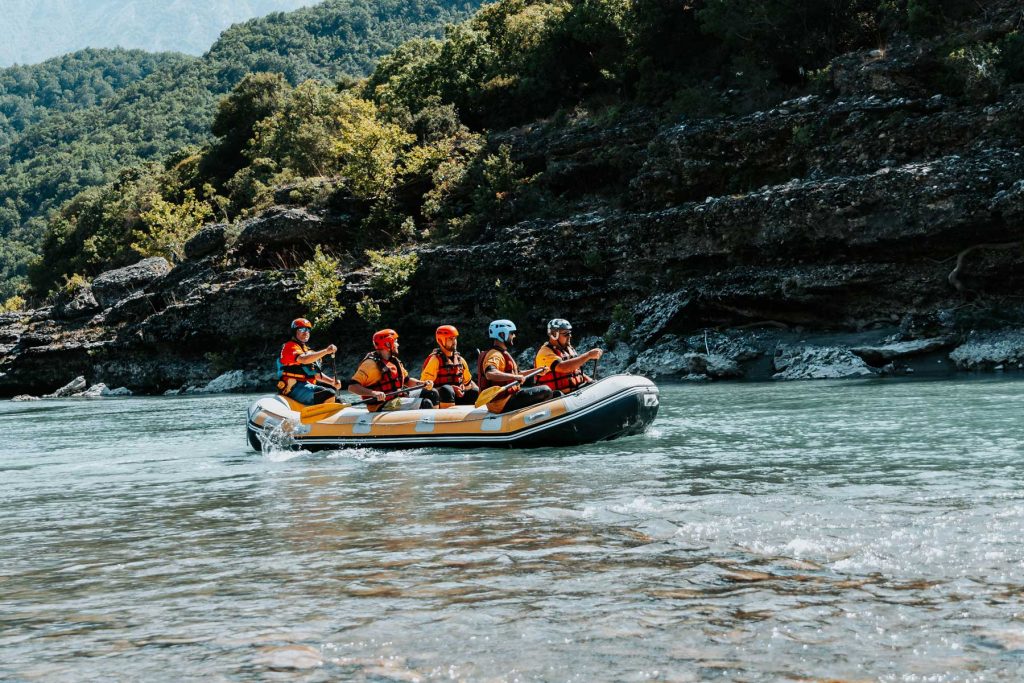 Zes mensen met helmen en reddingsvesten op varen op een rustige rivier, omringd door rotsachtig terrein en groene bomen, en genieten van een avontuurlijke roadtrip door de schilderachtige landschappen van Albanië – perfect voor moderne nomaden die op zoek zijn naar spanning in de natuur.