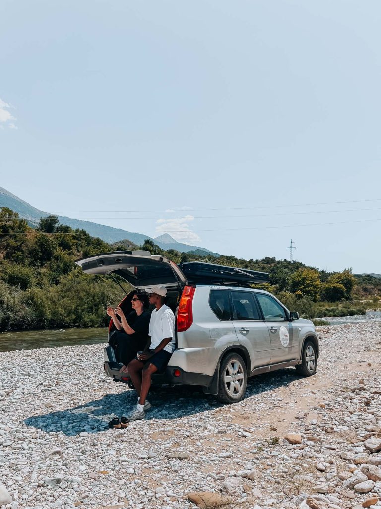 Twee mensen zitten in de open kofferbak van een SUV die op een rotsachtige rivieroever geparkeerd staat; ze genieten met volle teugen van hun roadtrip door Albanië, met heuvels en bomen op de achtergrond onder een heldere hemel.