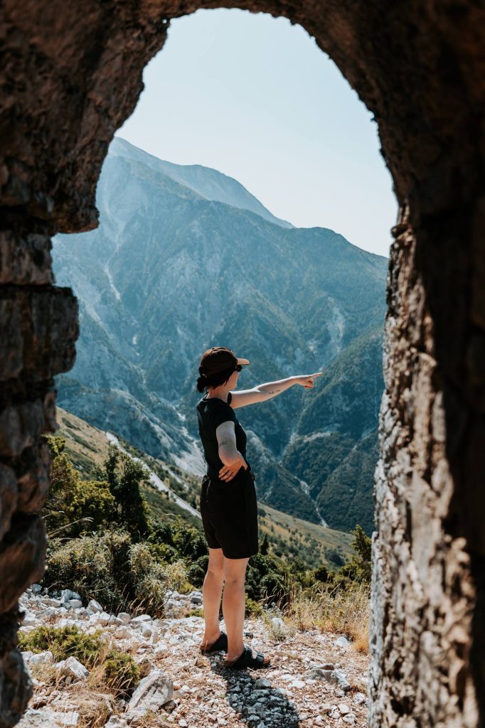 Een persoon in een zwarte outfit staat op een rotsachtige heuvel en wijst naar de bergen in de verte, met op de achtergrond een stenen boog – een tafereel dat de avontuurlijke geest van de Albanië Nomads weergeeft tijdens hun roadtrip door de woeste landschappen van Albanië.