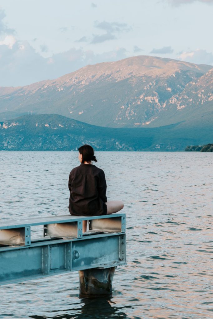 Iemand zit op de rand van een steiger met uitzicht op een rustig meer, met op de achtergrond bergen onder een heldere hemel, en legt zo de vredige sfeer van een roadtrip door Albanië vast.