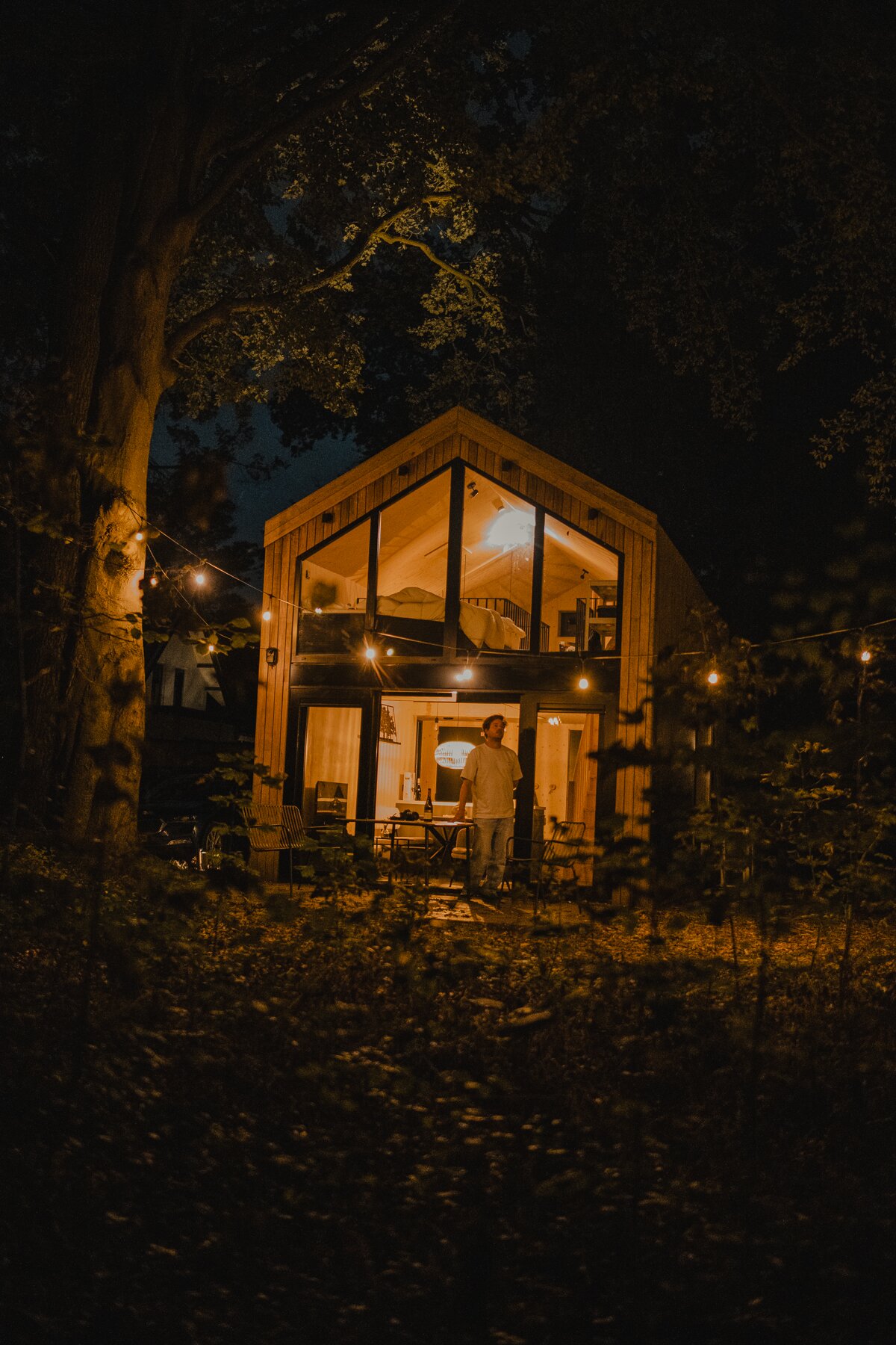 ’s Nachts staat er iemand buiten bij een moderne, met glas omgeven hut in het bos, verlicht door warme binnenverlichting en lichtslingers – een uitnodigend tafereel dat ‘het goede leven’ net buiten Breda perfect weergeeft.