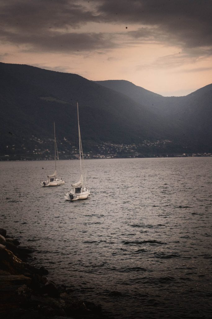 Twee zeilboten liggen voor anker op het Lago Maggiore bij Ascona, onder een donkere avondlucht met de bergen van Ticino op de achtergrond.