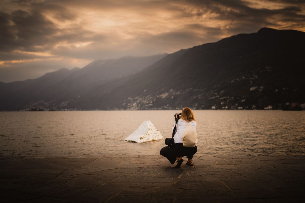 Een vrouw zit gehurkt aan de oever van het Lago Maggiore bij Ascona en fotografeert het meer in het laatste avondlicht.