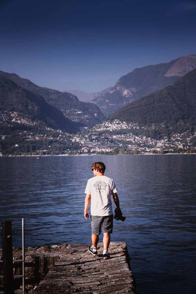 Een man staat op een stenen pier en kijkt uit over het blauwe Lago Maggiore, met de bergen en dorpen aan de overkant.