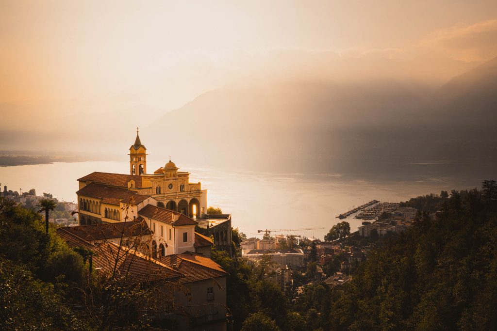 Het heiligdom Madonna del Sasso boven Locarno in het gouden ochtendlicht, met het Lago Maggiore en de bergen in de nevel.