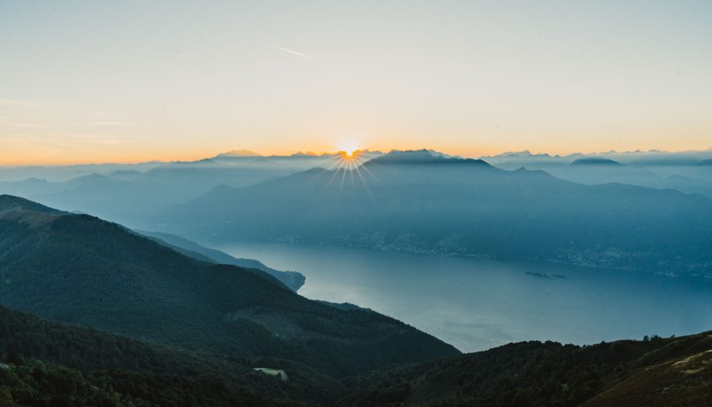 Het Lago Maggiore ligt in blauwe nevel tussen de bergen, gezien vanaf de Monte Gambarogno terwijl de zon achter de Alpen zakt.