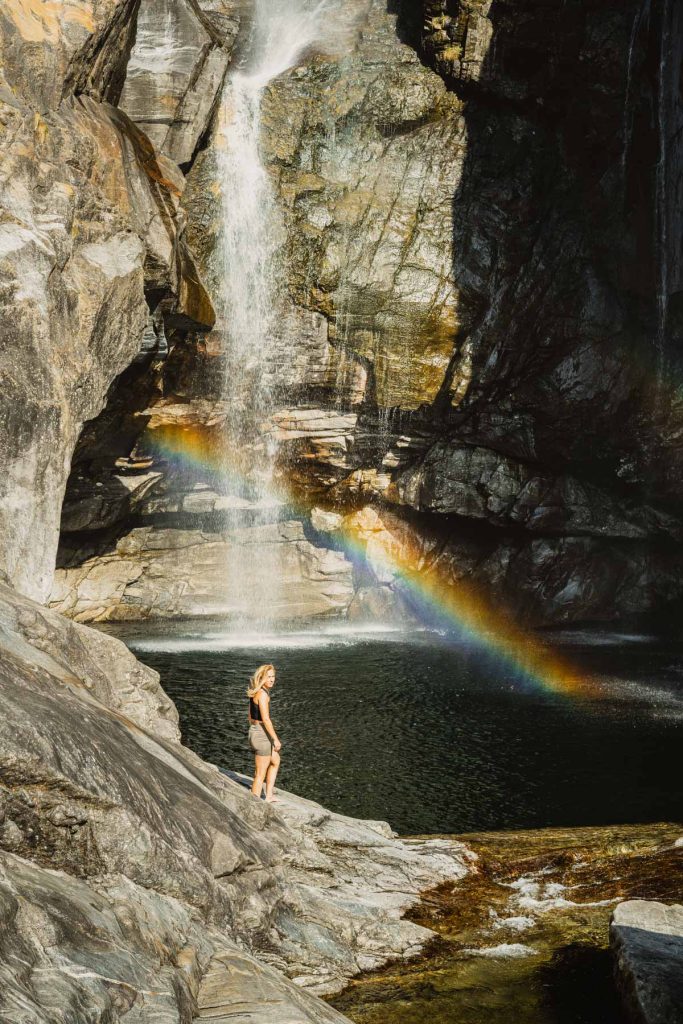 Iemand staat onderaan de Cascata del Salto in Maggia, terwijl er een regenboog in de nevel van de waterval hangt.