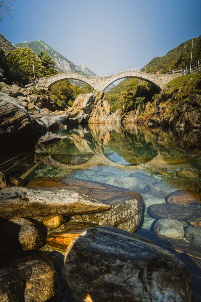 Door het heldere, groene water van de Verzasca zie je de stenen op de bodem liggen, met de Ponte dei Salti erachter.