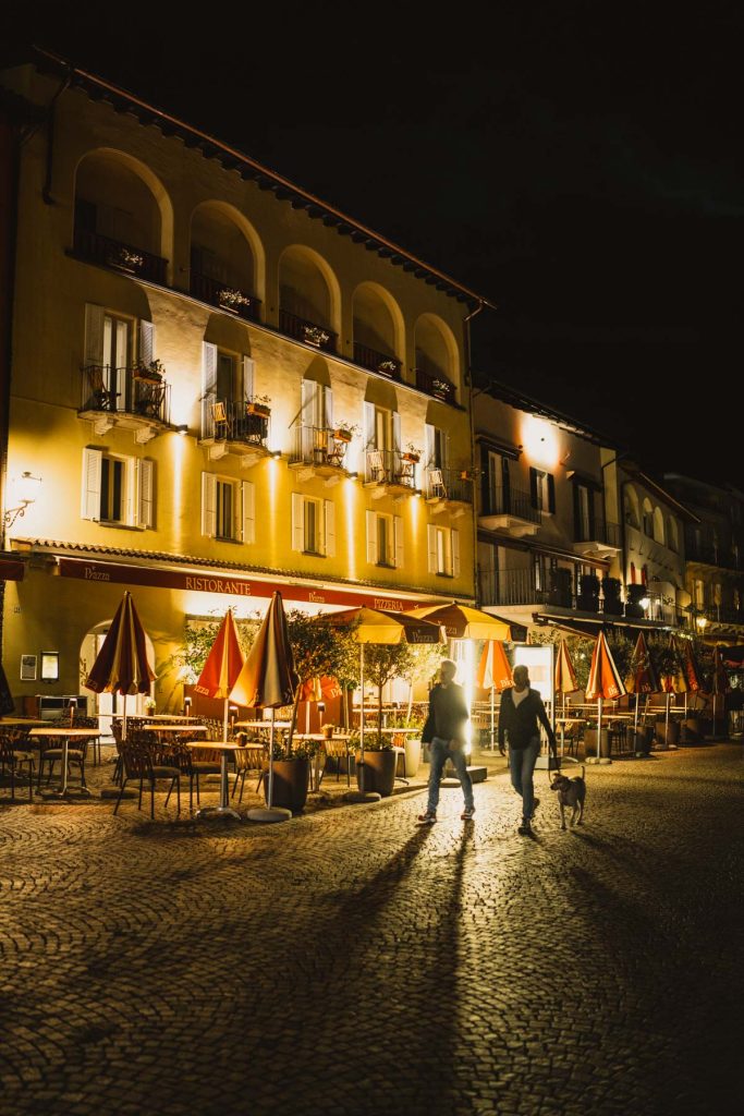 De arcaden aan de lungolago van Ascona baden 's avonds in warm licht, terwijl een paar mensen met een hond voorbij de terrassen wandelen.