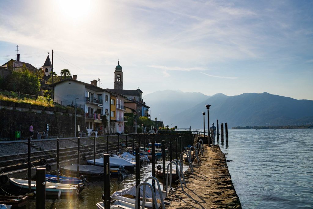 Bootjes liggen in een klein haventje aan het Lago Maggiore, met een dorp en een kerktoren in het zonlicht.